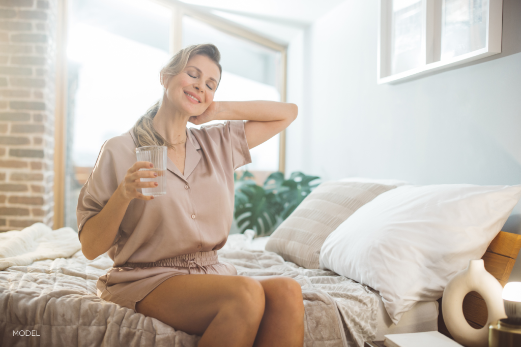 middle age woman sitting on bed, smiling and stretching while holding a glass of water